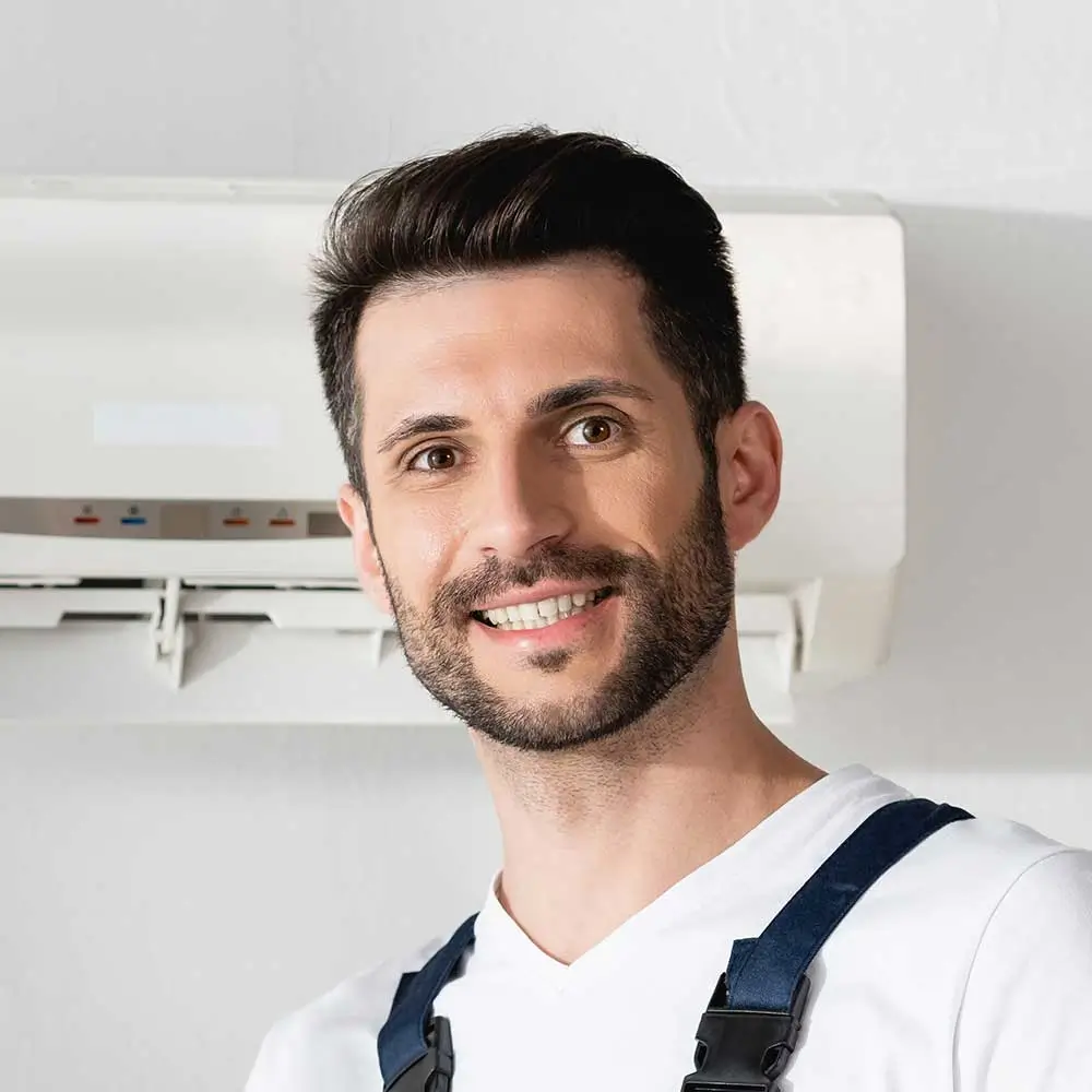 Man with a beard and mustache smiling in front of a white wall wearing a white t-shirt and blue suspenders.