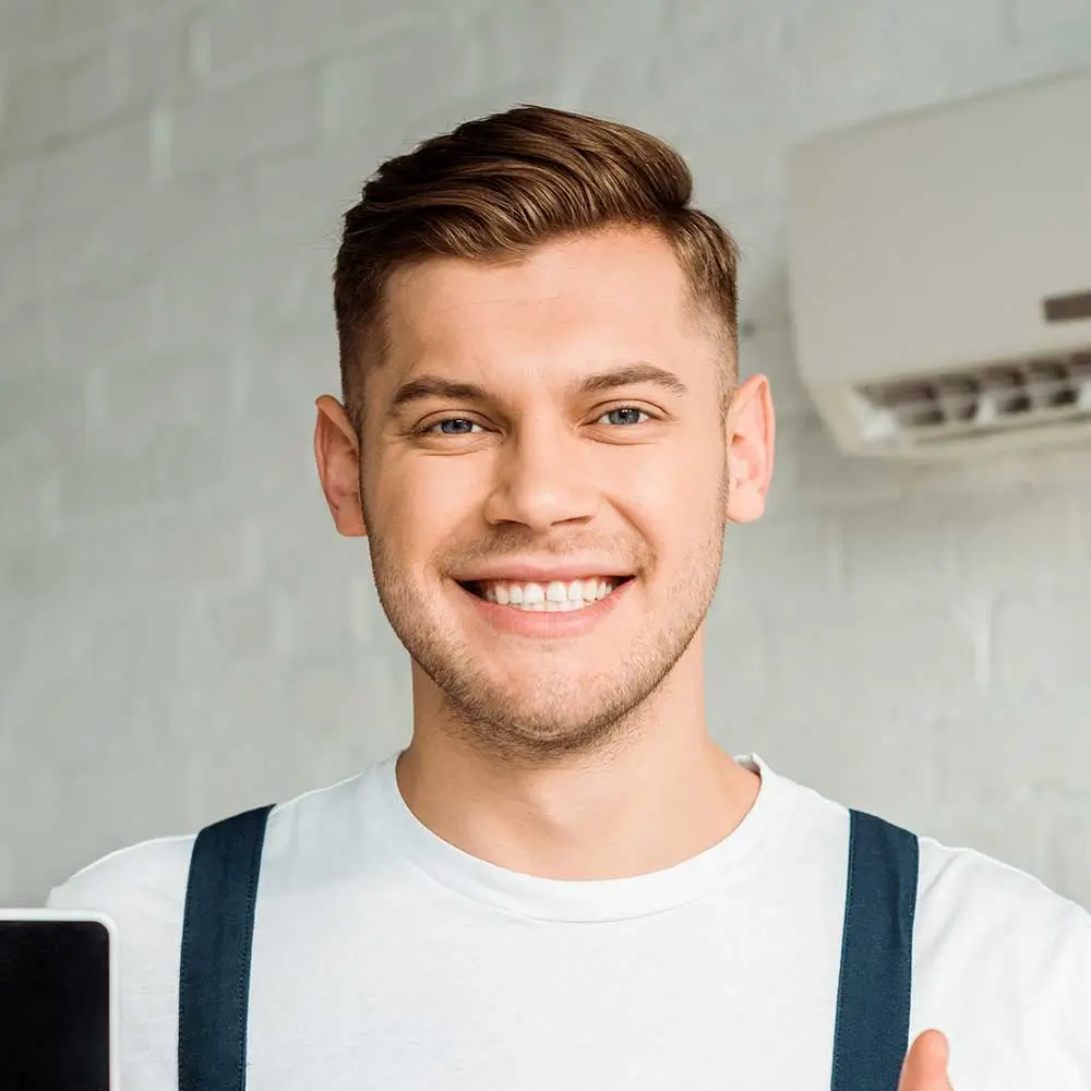 Young man with a cheerful expression wearing a white t-shirt and blue suspenders, holding a black tablet in front of a white brick wall.