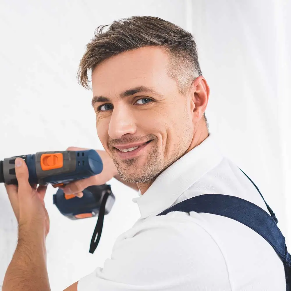Man smiling while holding a blue and orange power tool, wearing a white shirt and blue suspenders against a white background.