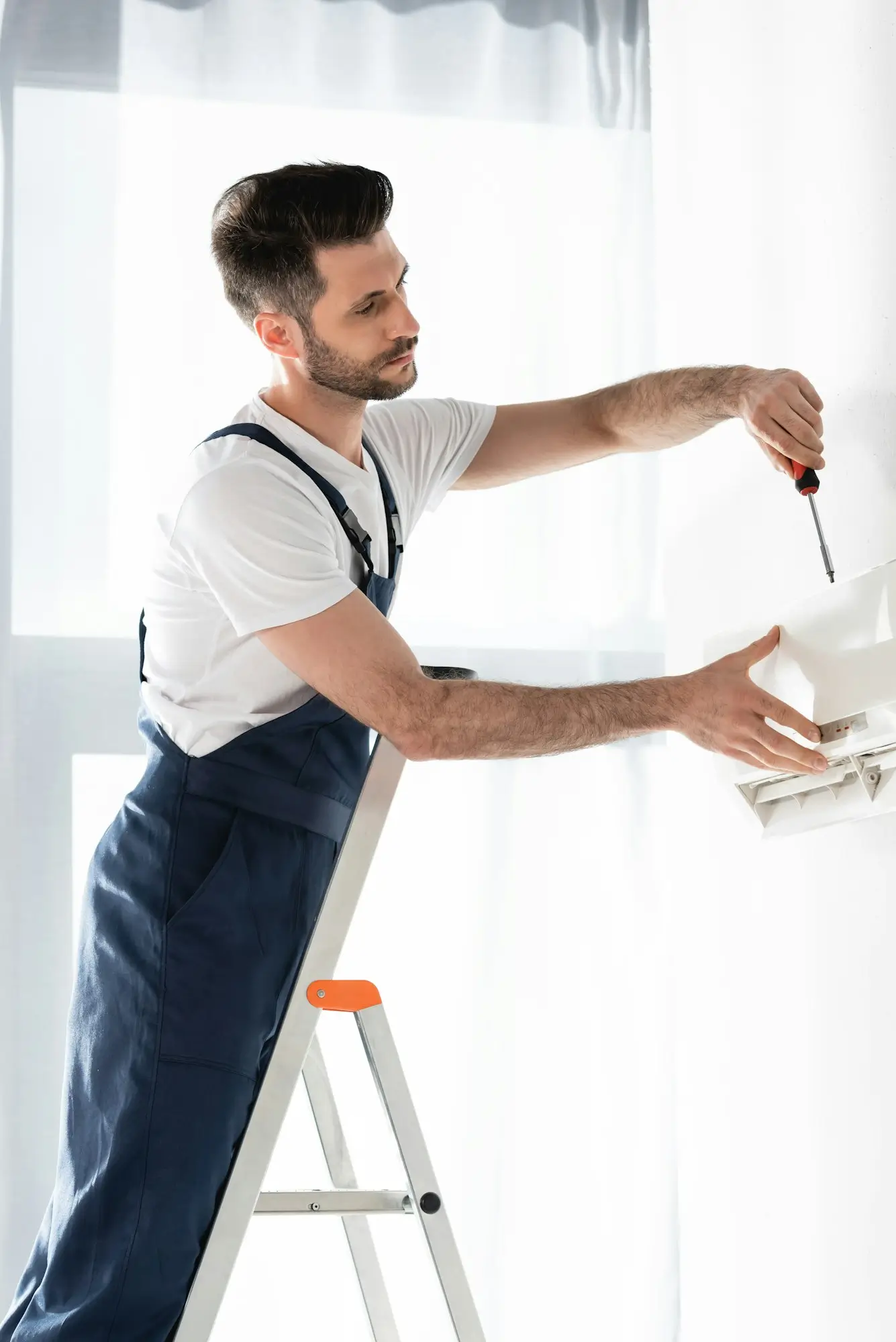Handsome repairman standing on stepladder and fixing air conditioner with screwdriver.